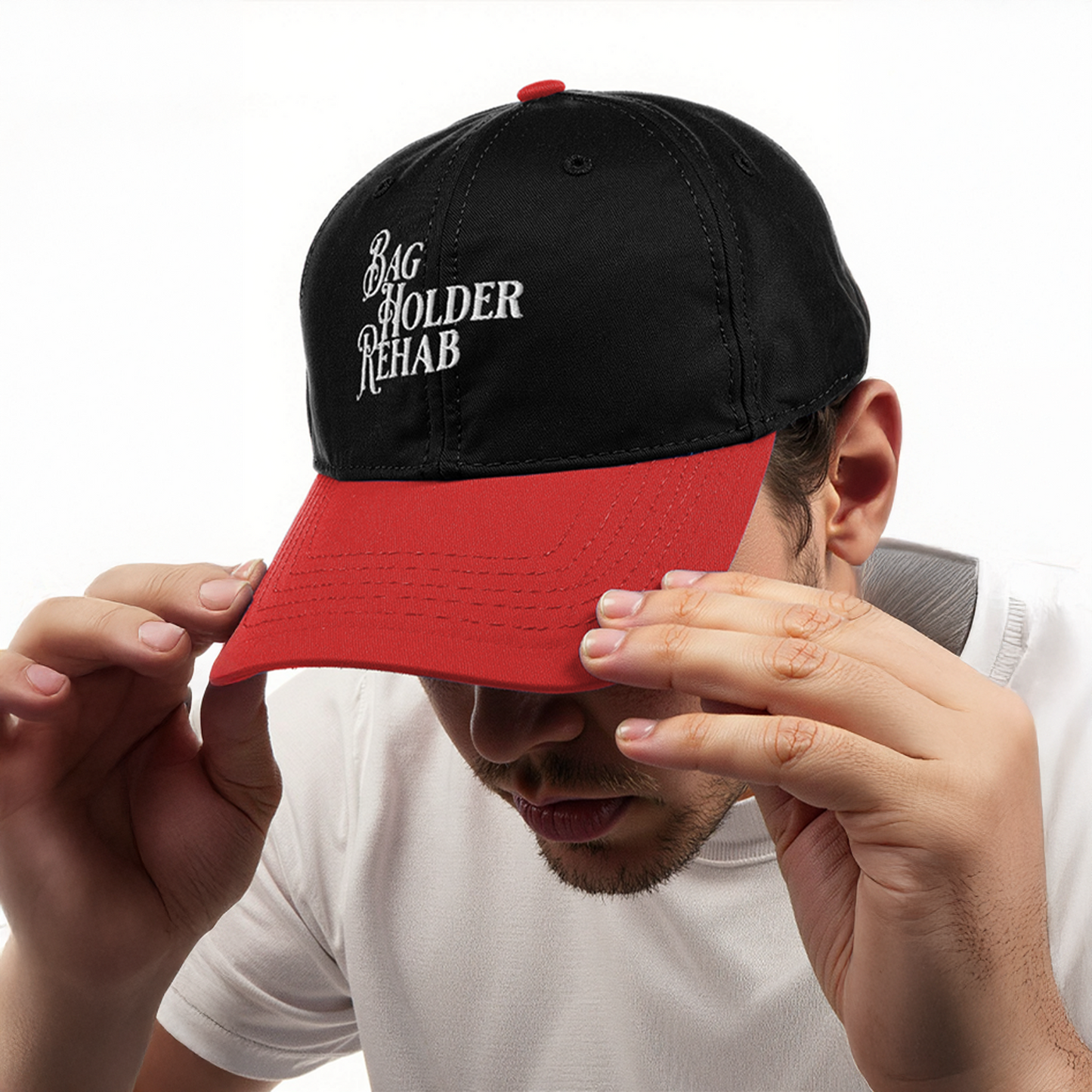 Man wearing black and red baseball cap with embroidered text “Bag Holder Rehab,” tilting brim downward while dressed in a white shirt