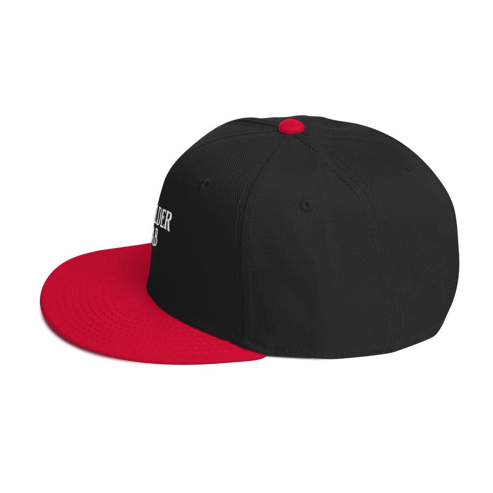 Man wearing black and red baseball cap with embroidered text “Bag Holder Rehab,” tilting brim downward while dressed in a white shirt