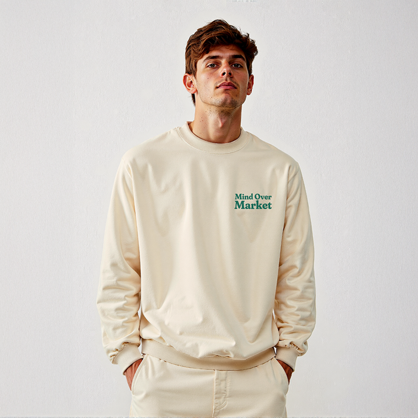 Man wearing cream “Mind Over Market” sweatshirt with hands in pockets, standing against a white background