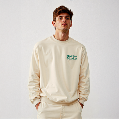 Man wearing cream “Mind Over Market” sweatshirt with hands in pockets, standing against a white background